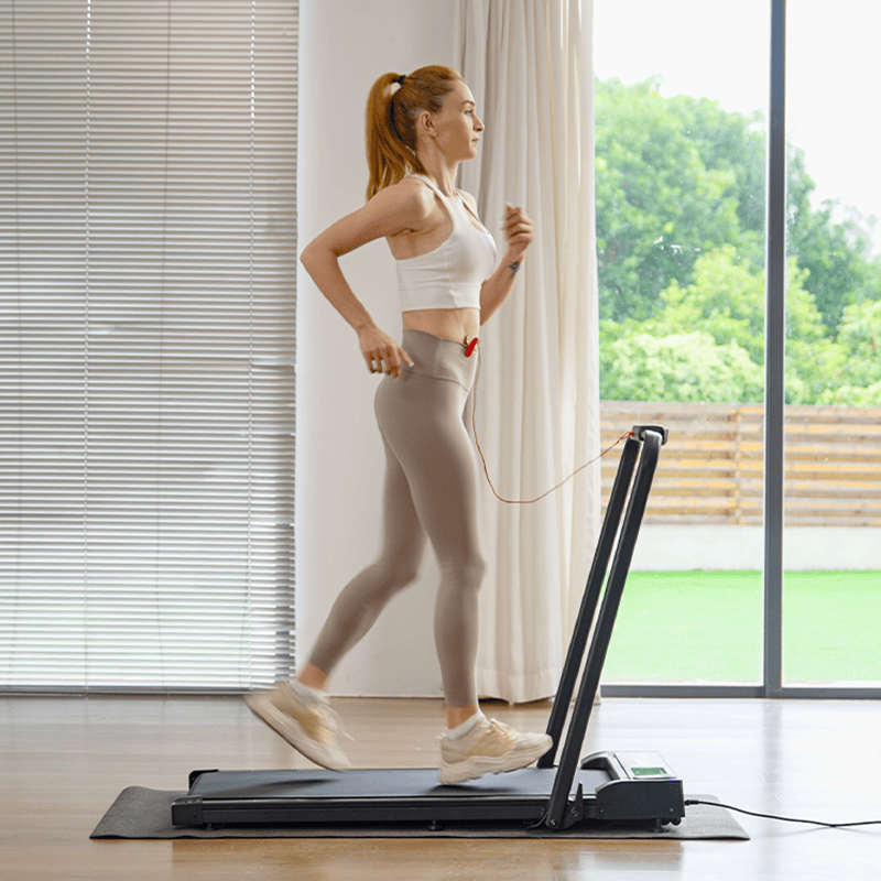 A woman is jogging on a treadmill JT51 indoors, wearing a white tank top and beige leggings. She has earphones in and a ponytail, with natural light streaming in from large windows that reveal a view of greenery outside.