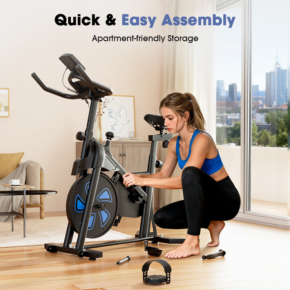 A woman in a blue sports top and black leggings assembles a stationary bike QK116C in a modern apartment with large windows and a city view.