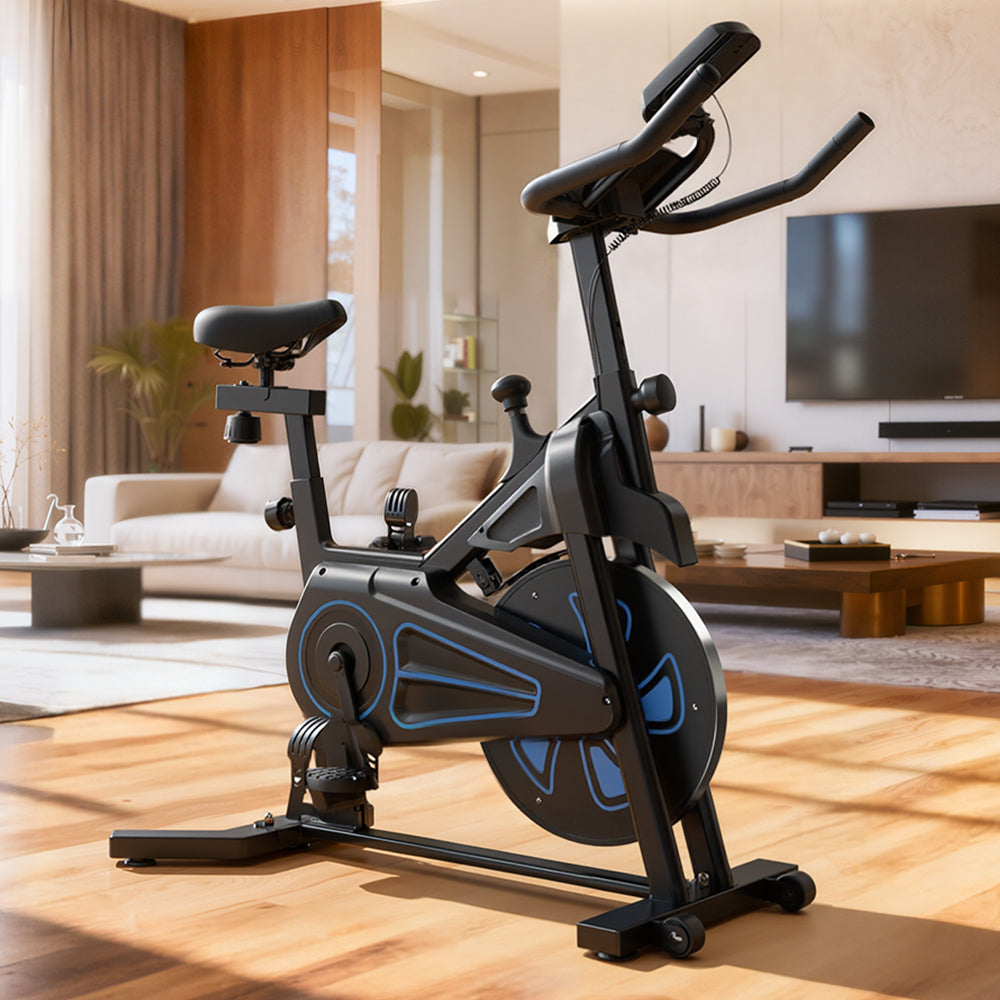 A sleek black exercise bike QK116C with blue accents is positioned on a wooden floor in a modern living room, featuring a large TV, a beige sofa, and natural light streaming through a window.